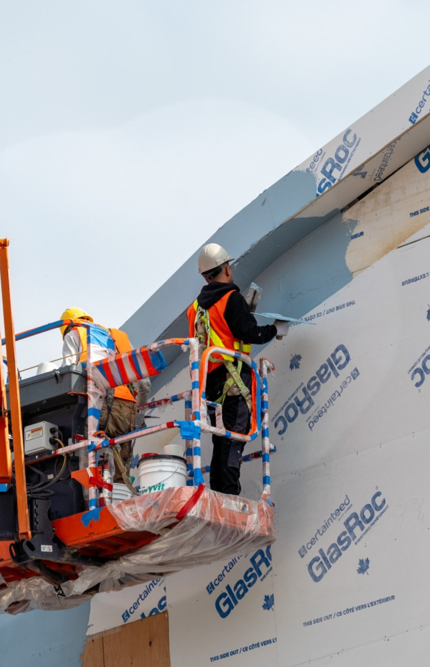 Two construction workers stand on a lift installing exterior cladding on a commercial building wall. Safety gear, tools, insulation panels, and a bucket are visible against a clear sky.